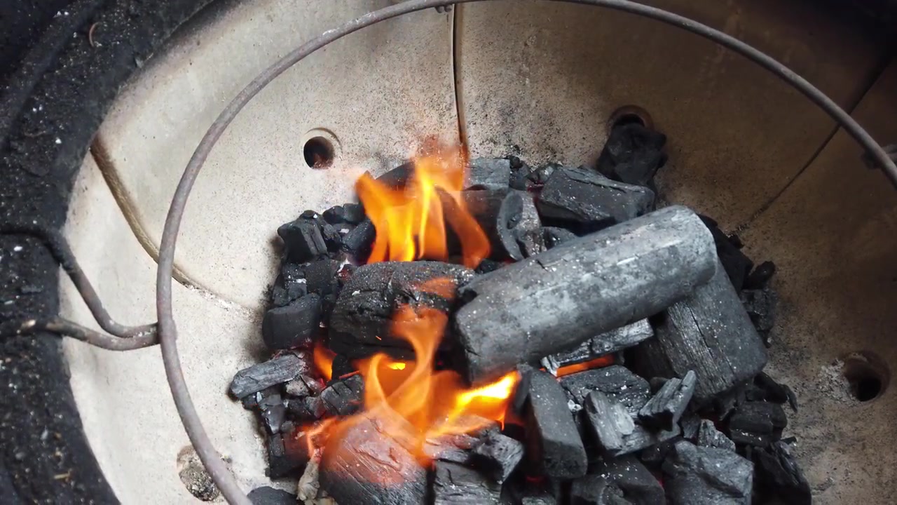Intenzivní oranžové plameny olizující černé uhelné brikety uvnitř kulatého keramického grilu, připravující se na grilování. Uhlíky jasně žhnou.