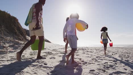 padres afroamericanos y sus hijos caminando con equipos de playa en la playa