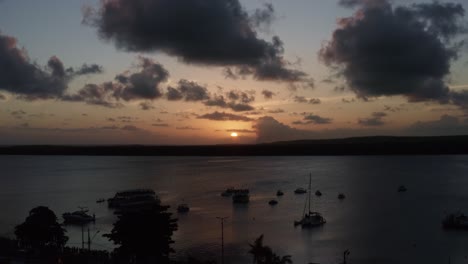 Trucking-left-aerial-shot-of-the-alligator-beach,-a-river-in-Cabedelo,-Paraiba,-Brazil,-near-the-coastal-capital,-Joao-Pessoa,-with-tour-boats-watching-the-sunset-as-a-man-plays-the-saxophone-below