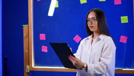 serious brunette lady in white shirt and glasses stands at glass wall with clipboard in hands. getting ready for presentation.