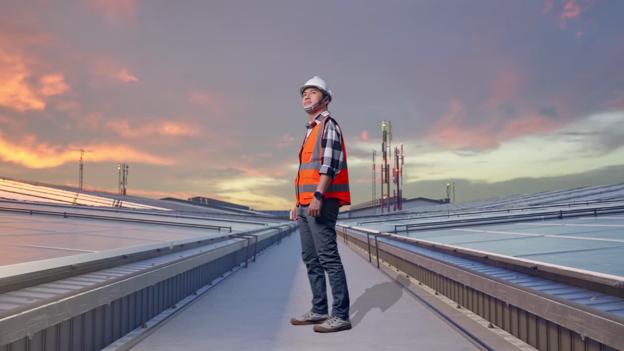 Vista lateral de cuerpo entero de ingeniero asiático con casco de seguridad mirando a su alrededor mientras está de pie e inspecciona paneles solares en la azotea.