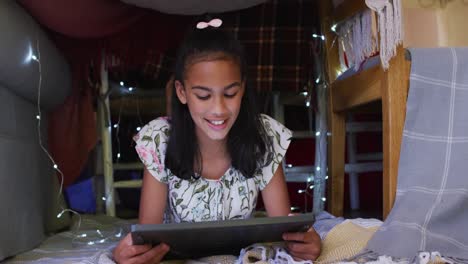 Happy-african-american-girl-lying-in-house-made-of-blankets,-using-tablet