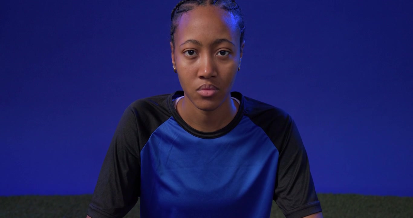 Inquadratura in salita, donna afroamericana in tenuta sportiva con un pallone da calcio, che si avvicina, allenandosi su un campo in erba sintetica.