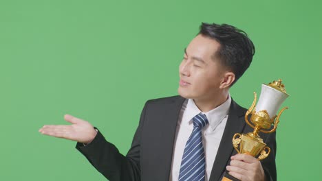 close up of asian business man in a suit and tie with a gold medal looking at a gold trophy in his hands, smiling, and pointing to side on green screen background in the studio