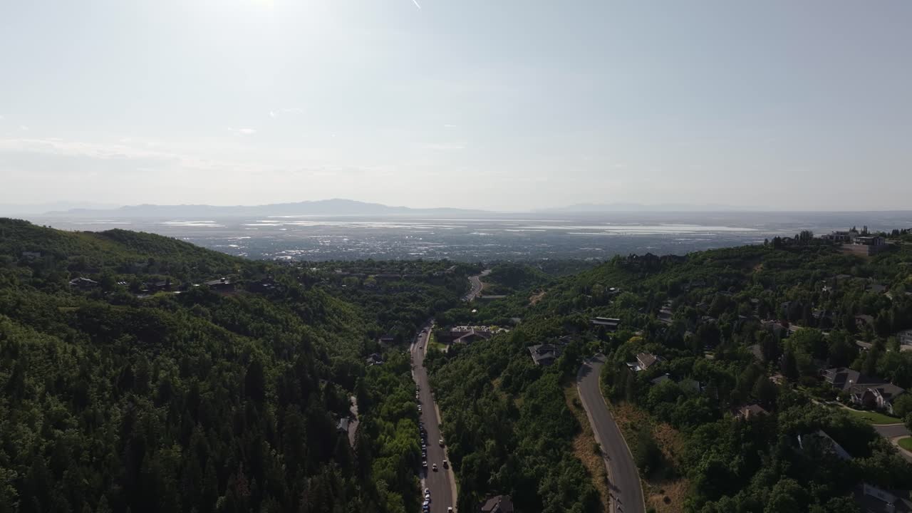 A wide aerial drone dolly in shot from Bountiful Canyon showing a road, forest, valley homes, cityscape, mountains, and the Great Salt Lake on a clear summer day