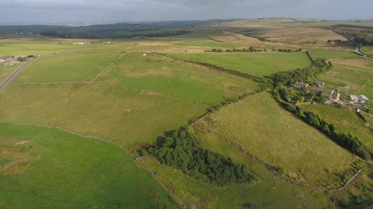 Sidewards drone footage at golden hour in the evening of rural West Yorkshire showing farmland, moors, farm buildings, country lanes, dry stone walls, sheep in fields, trees and towns in the distance