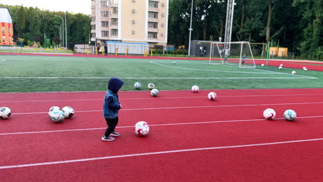 little kid in a hood walks by the red track in the stadium. baby boy walks up to the balls scattered around and picks one.