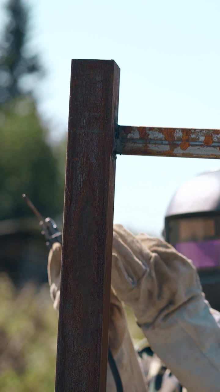 repairman in protective mask prepares metal parts of future fence for welding against clear sky on day
