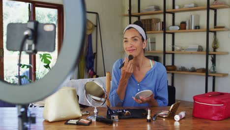 vlogger femenina de raza mixta aplicando maquillaje y grabando en el teléfono inteligente en casa