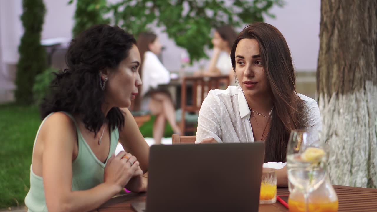 dos mujeres sentadas en la cafetería al aire libre - hablando, compartiendo ideas, mostrando algo en la pantalla de la computadora portátil, gestando, discutiendo