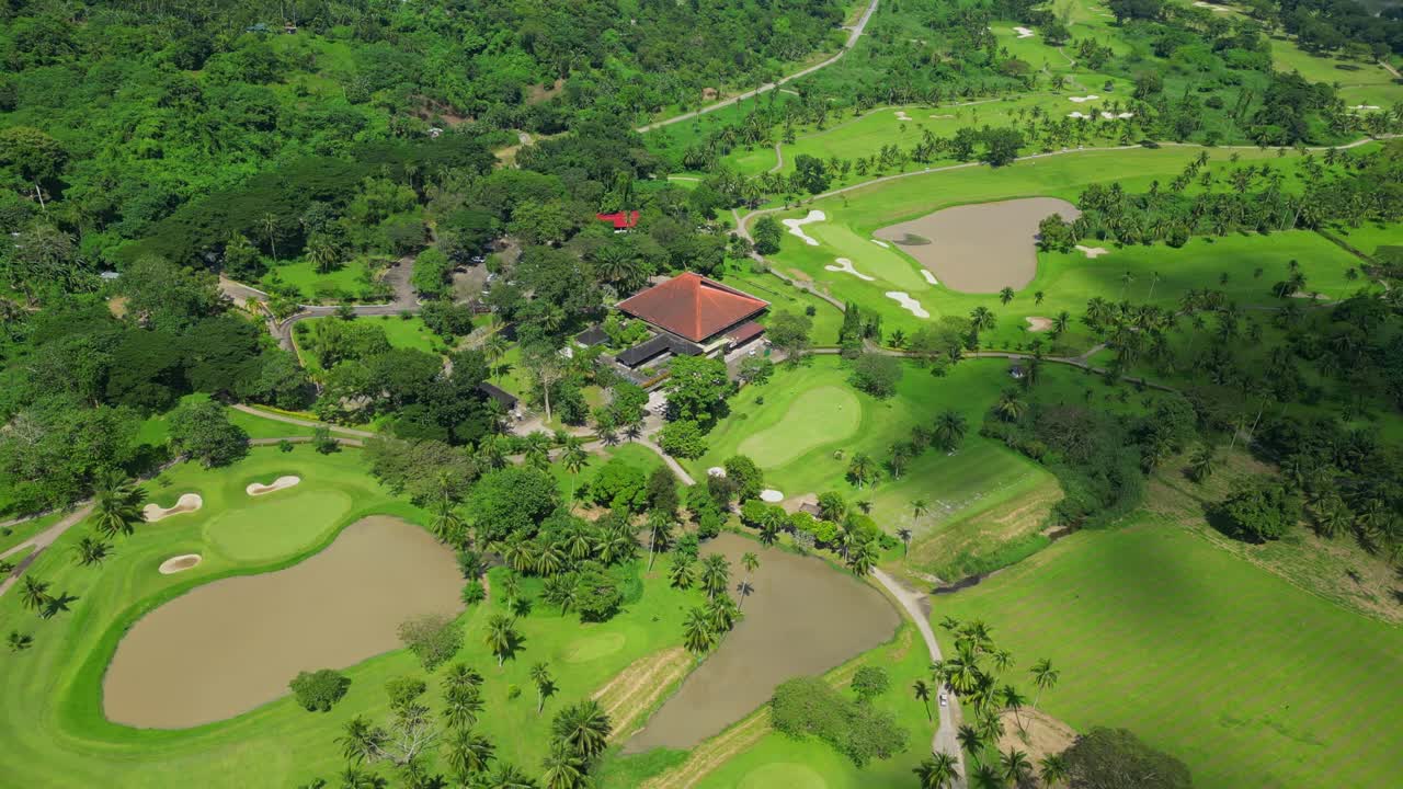A wider aerial shot of the country club showing the clubhouse, fairways, and water features with drifting clouds casting shadows in Calamba Laguna, Philippines
