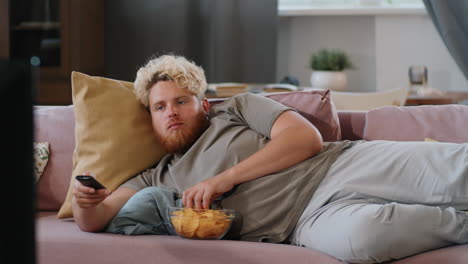 hombre con sobrepeso acostado en el sofá, viendo televisión y comiendo papas fritas en casa