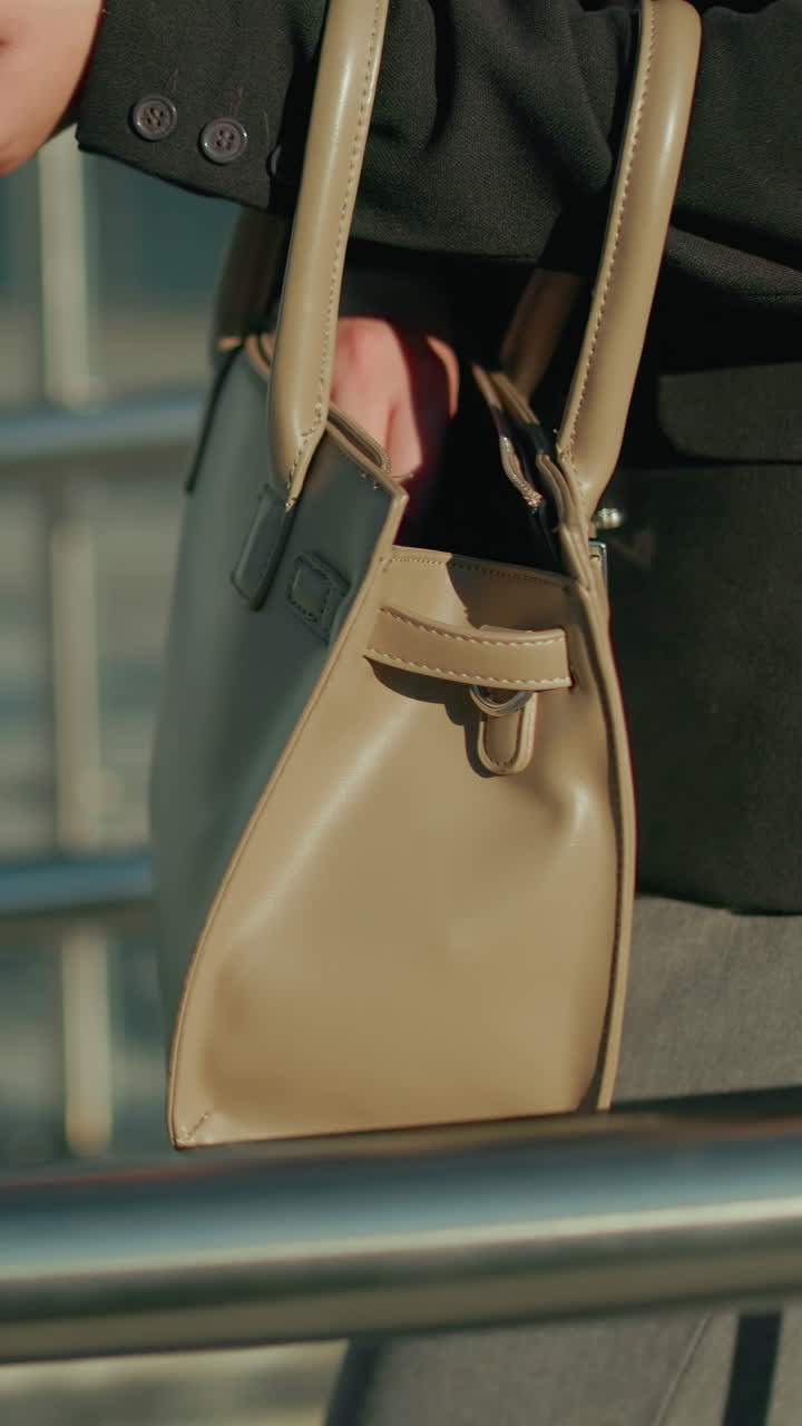 Close up side view of woman walking along pavement, checking handbag to retrieve keys, modern glass building , metal railings surround walkway, sunlight casts shadows in urban setting
