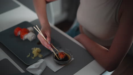 close up of woman in sleeveless top using chopsticks to dip sushi into soy sauce at table, enjoying casual indoor meal with visible plate of assorted rolls, ginger slices, and napkin nearby