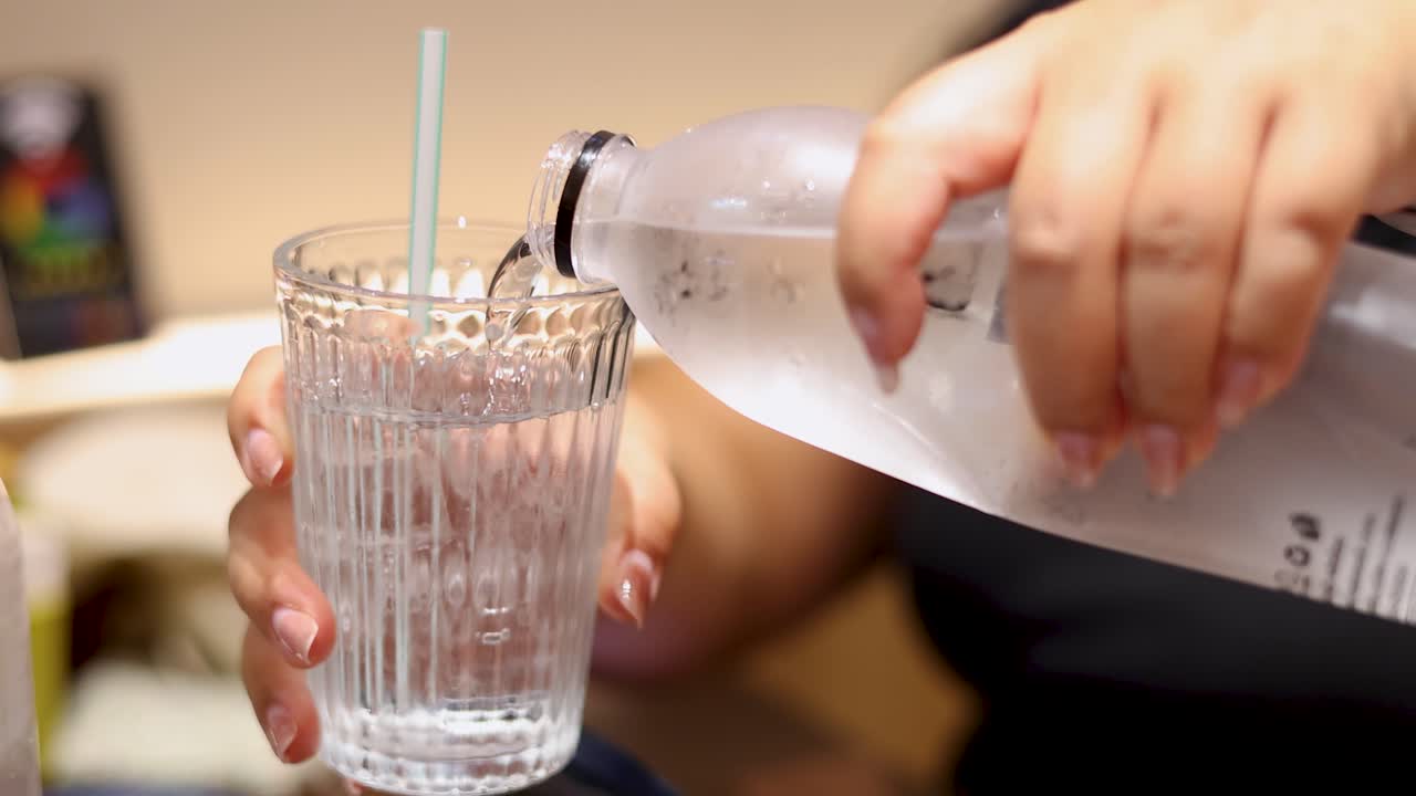 A person pours clear water from a plastic bottle into a textured glass with a straw, under warm indoor lighting, using a close-up perspective