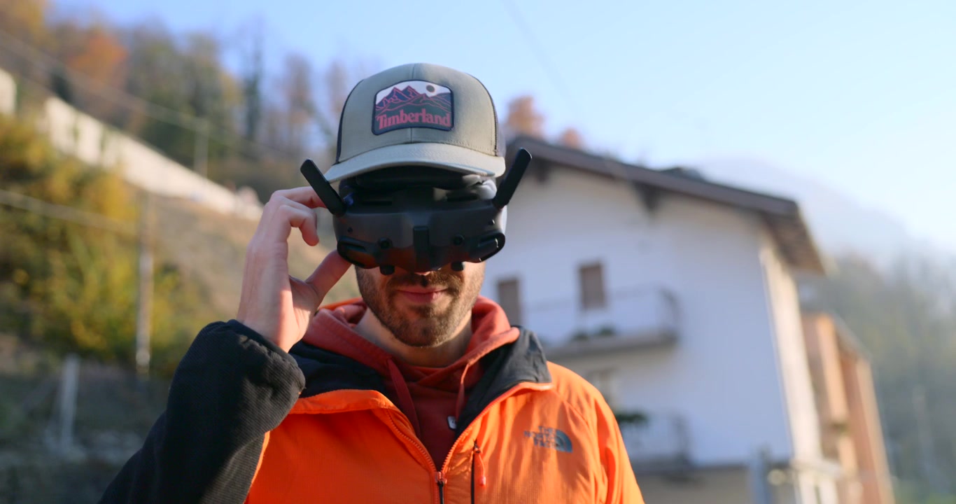 Un uomo regola gli occhiali FPV del drone prima del volo in un villaggio di montagna, impostando il dispositivo per un controllo aereo immersivo e un'avventura all'aperto.