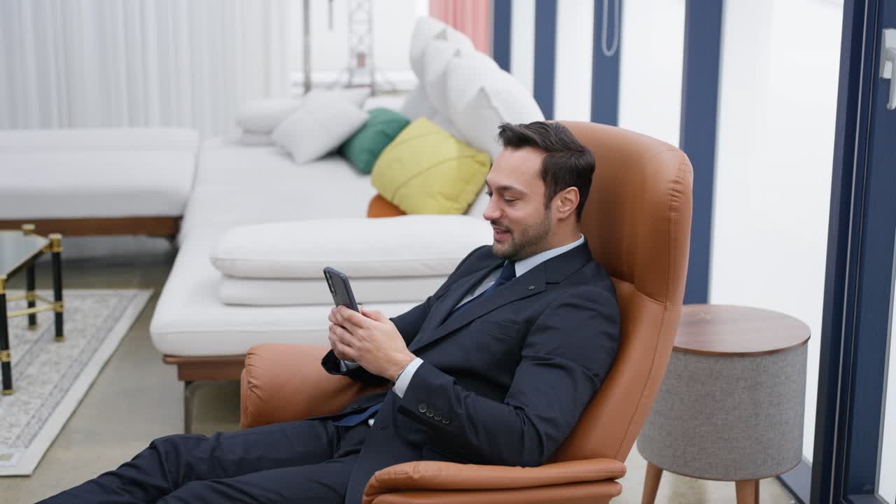 European businessman enjoying success win on mobile phone on recliner armchair at home. Male reading good news on smartphone, showing yes gesture. Surprised man celebrating victory on phone saying wow