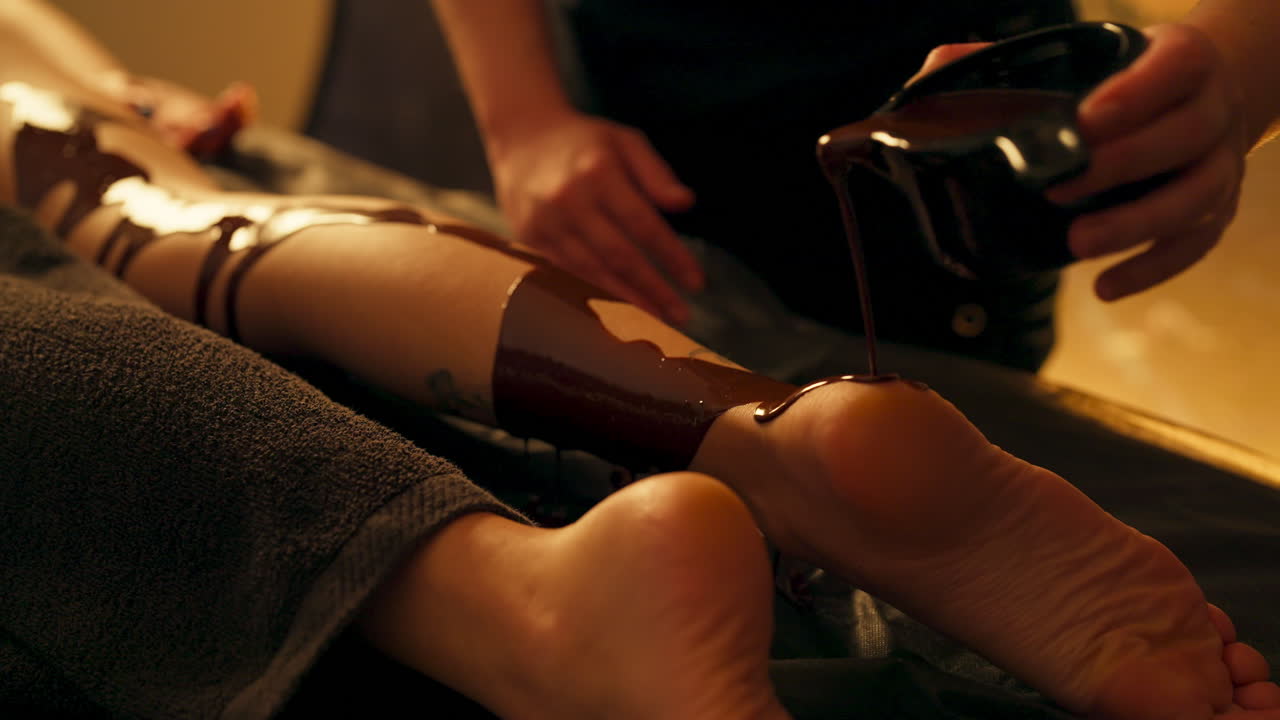 Therapist Pouring Melted Chocolate Onto Leg Of Female Client. Chocolate Massage At Spa. closeup shot