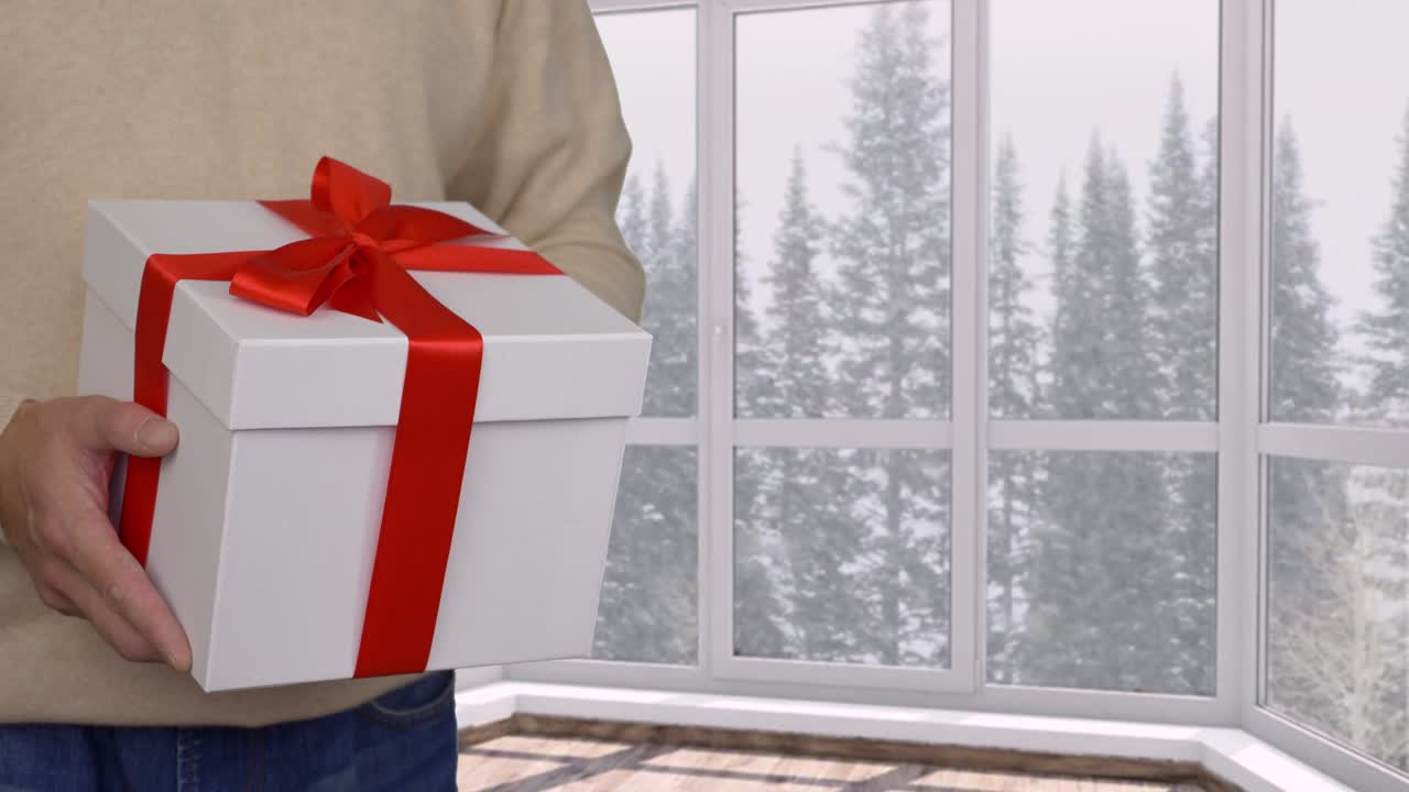Man presenting gift box with red ribbon on christmas or new year, snowfall outside the window