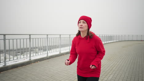 woman in red beanie and hoodie jogging near iron railing on foggy pathway, distant bridge visible across railing with cars moving, capturing active fitness lifestyle and urban winter environment