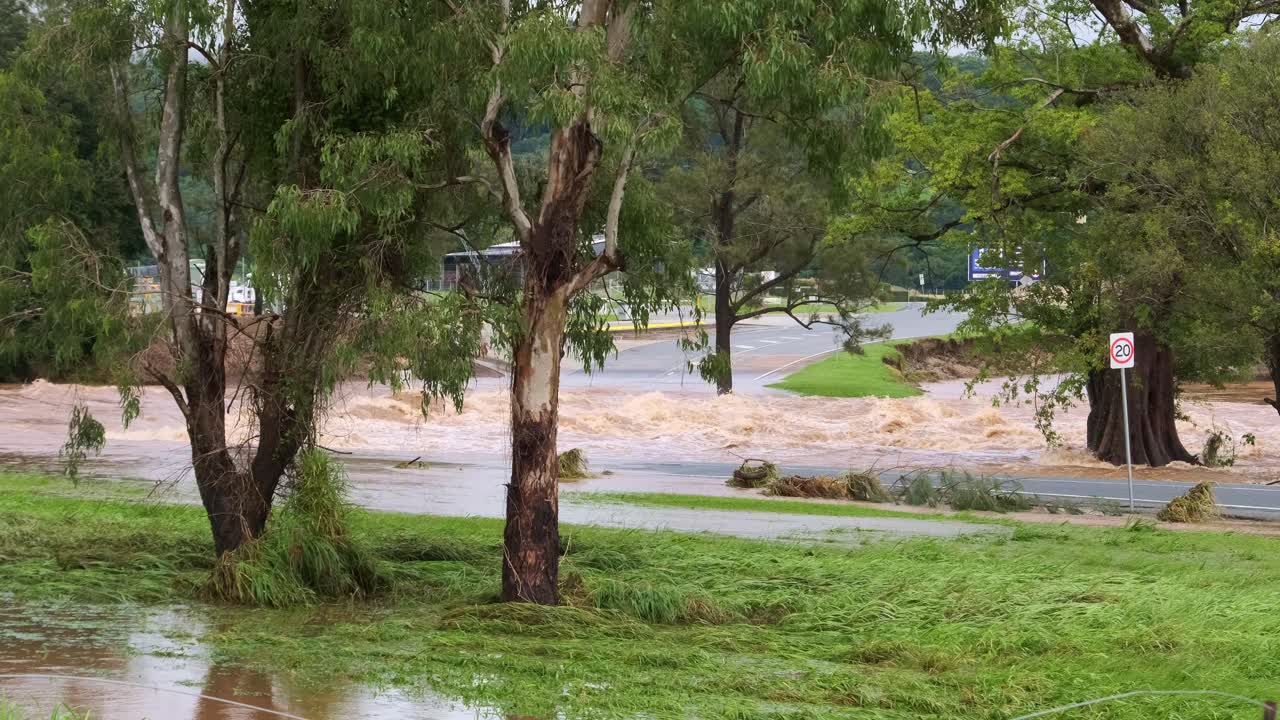 A flooded river overflowing its banks on the Gold Coast in Queensland, Australia. Brown water flows rapidly through green grass and trees, creating a dramatic natural scene.