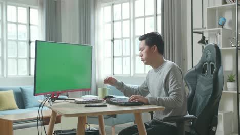 asian man in long sleeved t-shirt and black pants being tired while using green screen desktop computer for working at home.