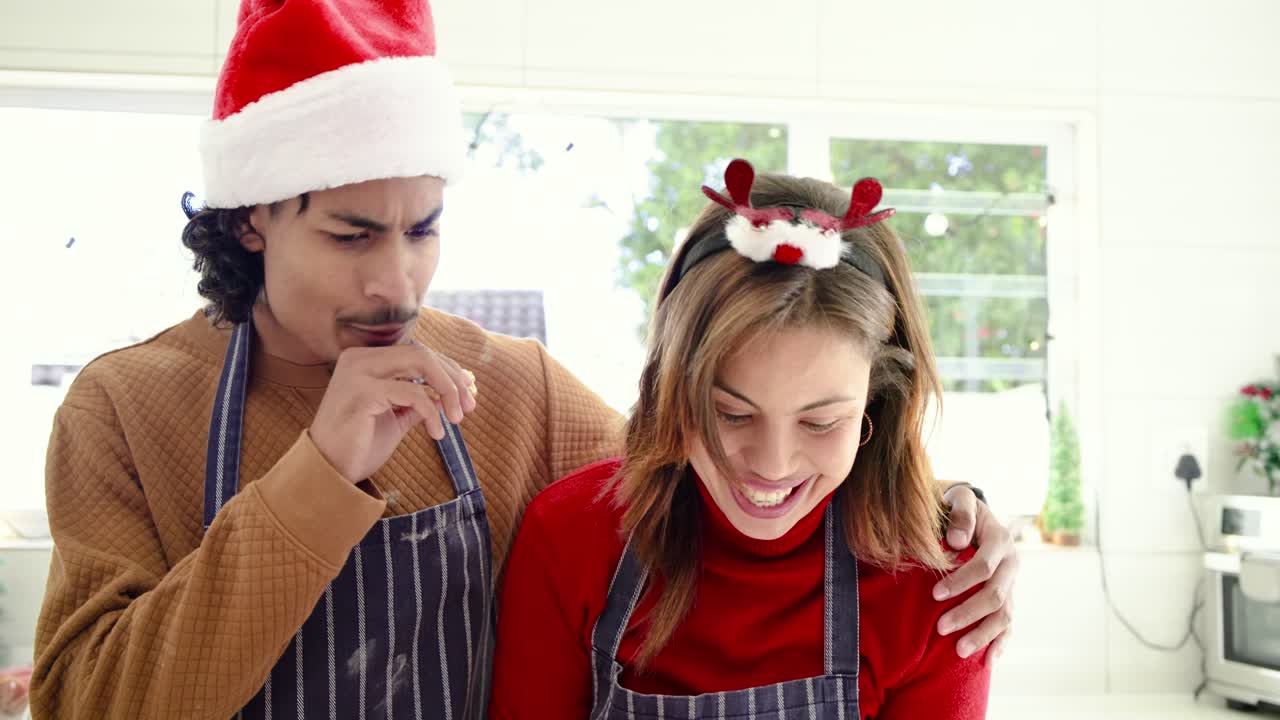 Diverse couple wearing Santa hats spotting cookies on rack, icing treats, tasting flavor in kitchen