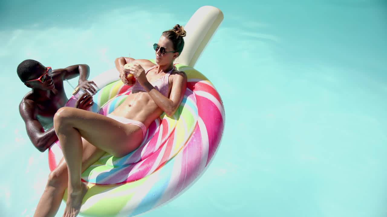 Relaxing in pool, diverse couple enjoying drinks on colorful inflatable float