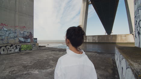 Medium close-up shot of a young woman contemplating the urban, graffiti filled scenery. Strong winds under the Vasco da Gama bridge. Urban isolation. Wonder and awe in solitude