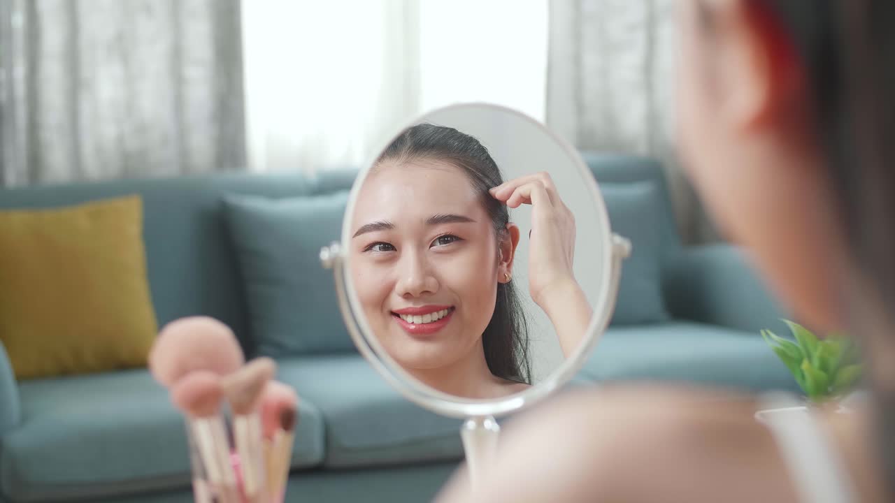 거울에 반사 아름다운 젊은 아시아 여자가 미소 짓고 머리카락을 고치고 집에서 메이크업하는 동안 준비하고 있습니다.