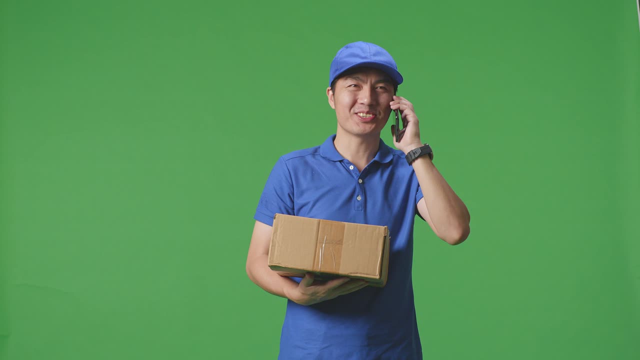 푸른 유니폼을 입은 아시아 남성 택배기사 가 스튜디오 에서 녹색 스크린 배경 에 박스 를 배달 하는 동안 스마트폰 으로 이야기 하고 있다