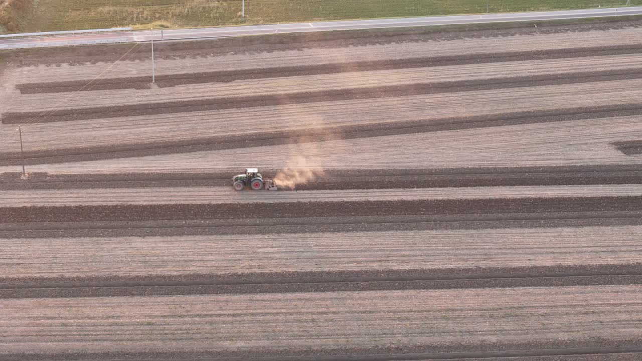 Tractor plowing structured rows in a dry Po Valley field near Piacenza, Italy, with dust plume, utility poles, and access road, drone panning