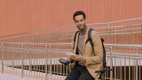 young american man in formal clothes using a tablet while leaning on bike in front of a prefab metal building 1