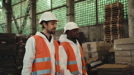 A-brunette-man-with-a-beard-in-a-white-uniform-and-an-orange-protective-vest-walks-with-his-Black-skinned-male-colleague-and-communicates-at-a-large-waste-processing-and-sorting-plant