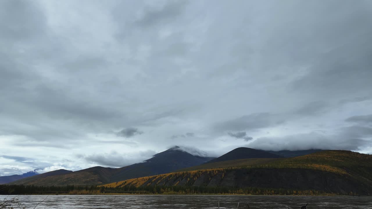 Witness the dynamic transformation of clouds as they drift over majestic mountains and a flowing river. This captivating display showcases the beauty of nature throughout the seasons.
