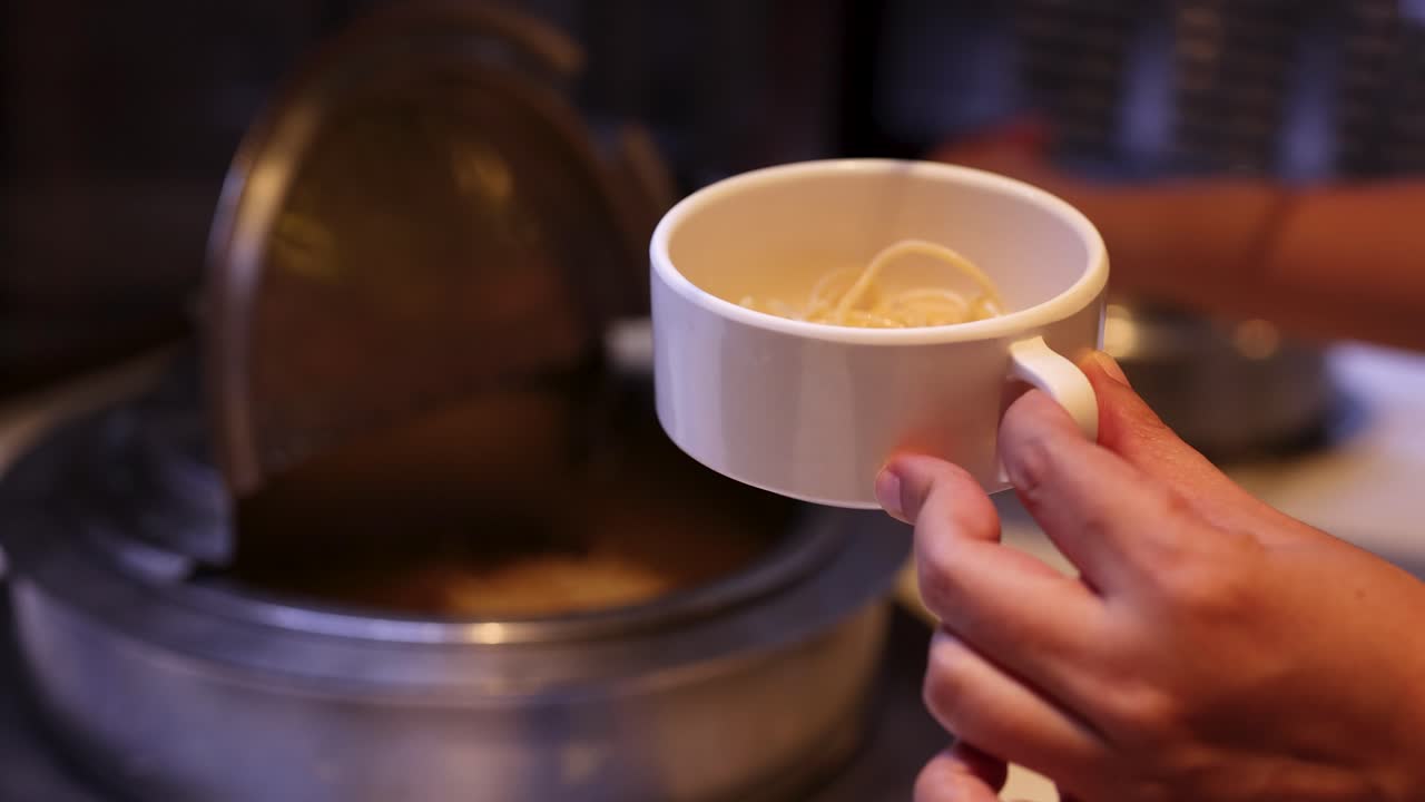 Hand ladles steaming broth over noodles into cup at buffet soup station, warm indoor lighting