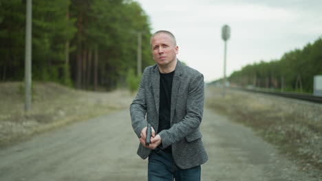 a close view of a man in a gray jacket and jeans walking along a dirt path beside a railway track, holding a handgun aiming with it, with a blurred view of a forest and distant power lines