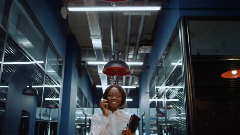 cheerful afro woman using phone in corridor. woman talking on phone in office