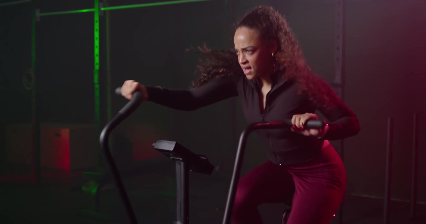 Donna afroamericana sente il timer, pedala su una cyclette ad aria in una palestra in un magazzino, indossando abiti da allenamento.