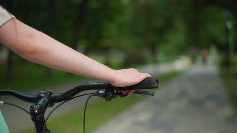 close-up of person s hand gripping bicycle handlebar while strolling along a park pathway, surrounded by blurred greenery and an out-of-focus figure in background