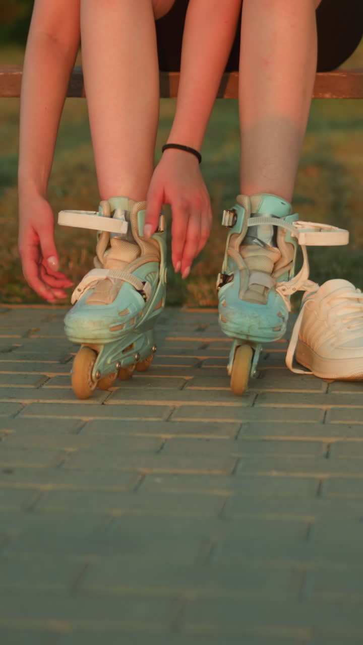 Lady removing strap of left leg roller skate while seated on bench in park with two white sneakers by her side, with blurred background featuring greenery