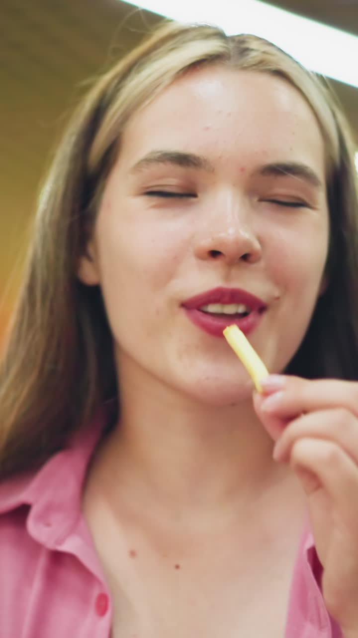 Woman seated in restaurant takes a potato chip, smiles, takes a bite, closes eyes in satisfaction, and shakes head, savoring the delicious taste, with bright restaurant lights in the background
