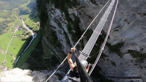 a pov shot from above of a man walking over a small and narrow hanging bridge in via ferrata with village murren below in the green and lush valley