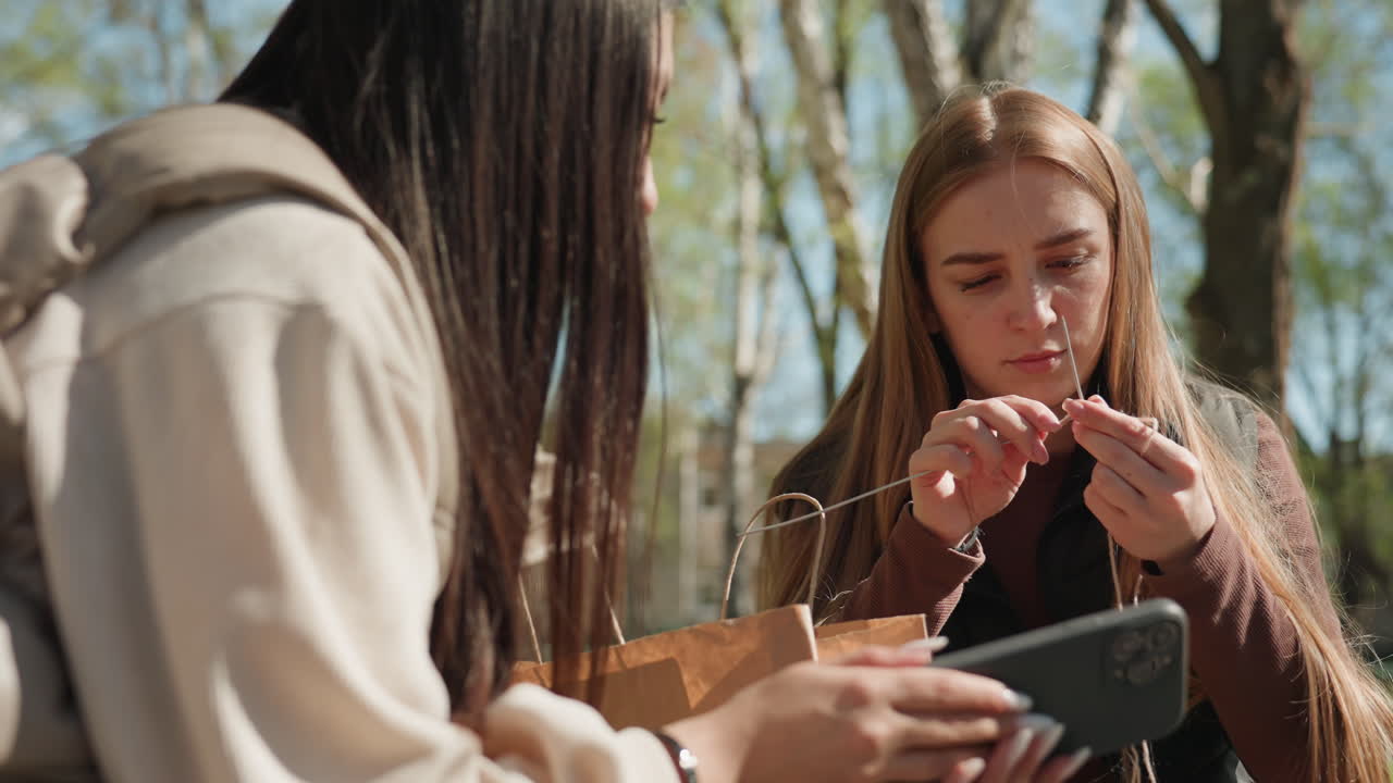함께 밖에서 물건을 구경하는 여성들, 편안한 공원 나들이 중에 최근 구매한 상품을 살펴보는 여성들, 여유로운 야외 공원 나들이 중에 쇼핑백을 확인하는 여성 친구들