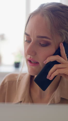 freelance woman with fair hair bun talks on phone working online via modern laptop against bright window in comfortable home room slow motion closeup