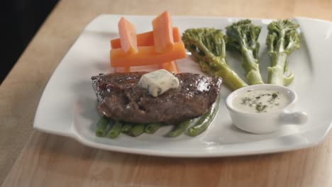 medium slider shot of a succulent steak with butter on top and sides of carrots broccoli and green beans on a wooden kitchen table