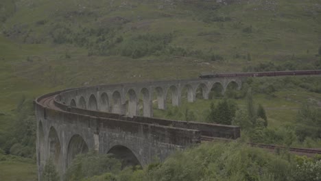 famous steam train crossing a curve arched stone bridge in a beautiful landscape in scotland uk. green landscape and white smoke