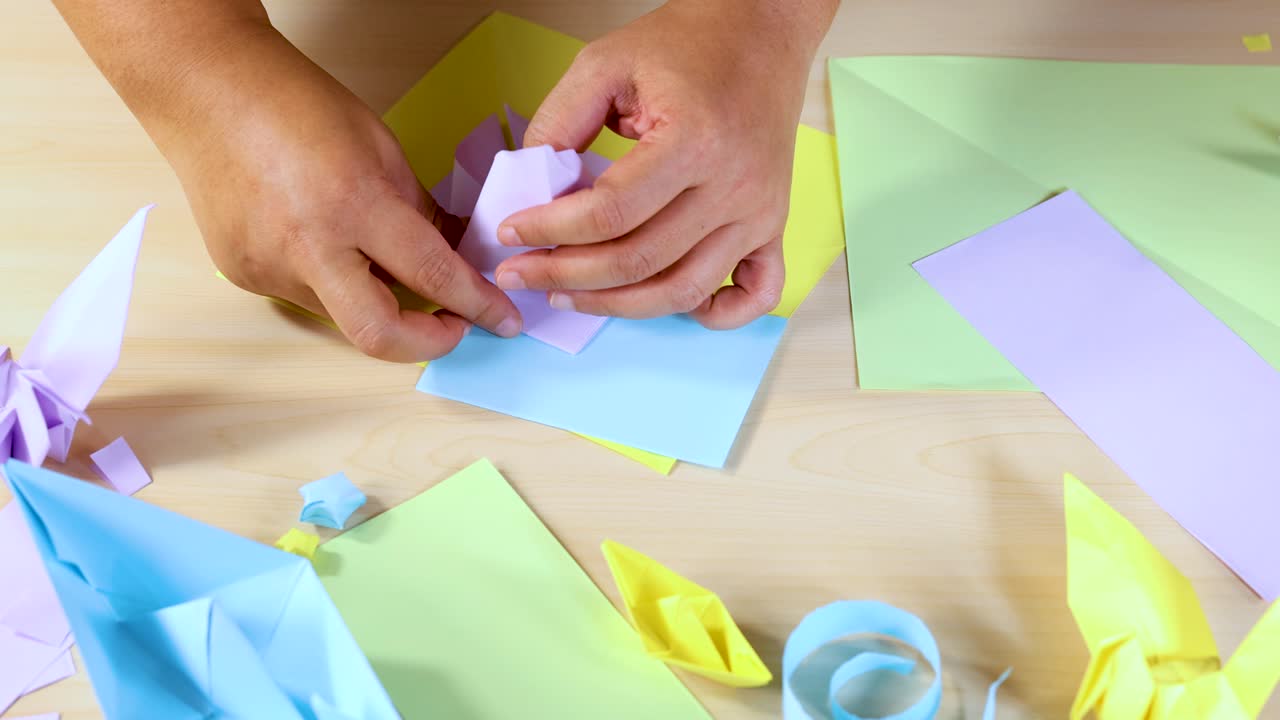 Person folds pastel origami paper, surrounded by finished crafts, under bright, even lighting, overhead view