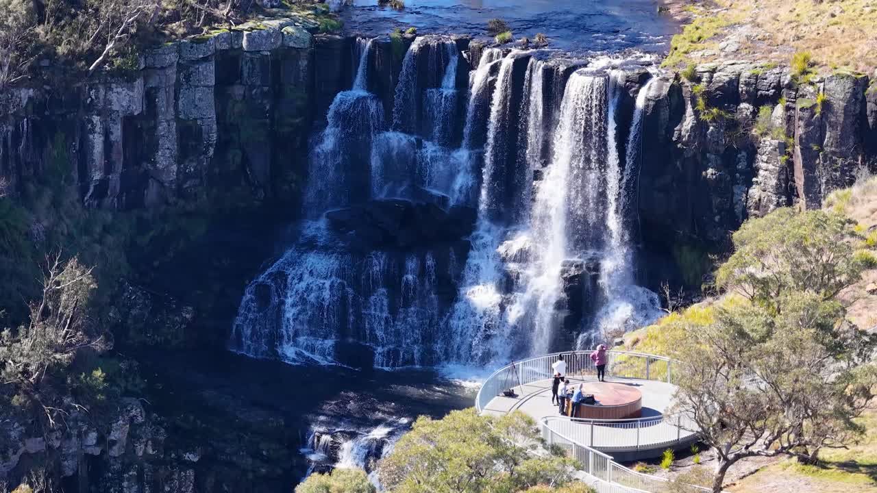 Aerial camera glides over a scenic lookout platform above a multi-tiered waterfall, surrounded by lush trees and rocky cliffs in bright daylight