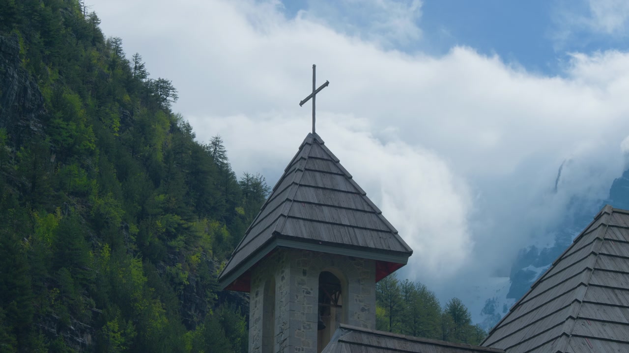 Campanile di chiesa in mattoni con sfondo di montagne alpine e cielo blu, scatto statico da treppiede. Primo piano della cima di una storica cappella di montagna con nuvole